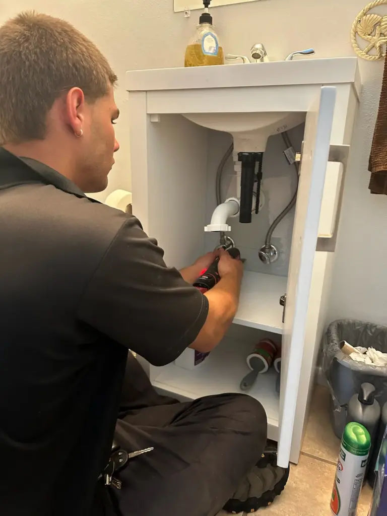 Technician kneeling at an open bathroom vanity, working on the supply lines under the sink with a tool.
