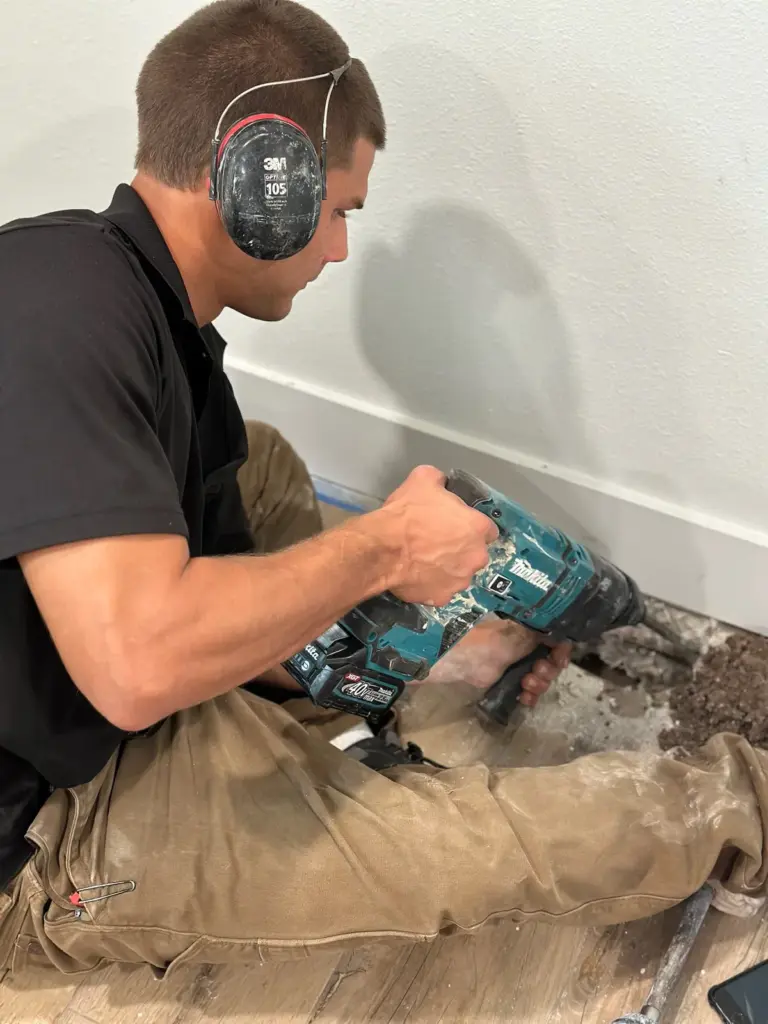 Technician wearing ear protection kneeling and drilling into the floor at a wall with a rotary hammer.