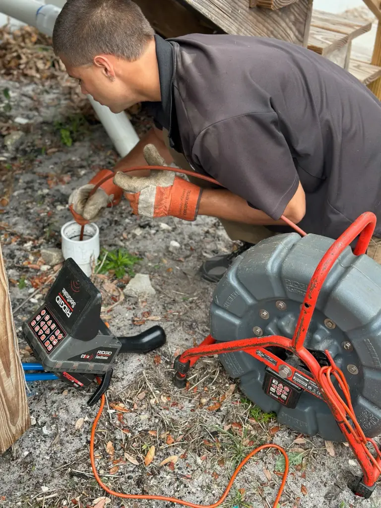 Technician crouched feeding a camera cable into a white cleanout pipe, with the red reel and monitor beside him.