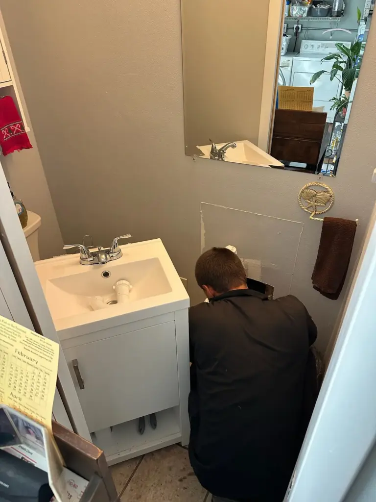 Technician kneels beside a white bathroom vanity, working at an access opening cut in the wall.