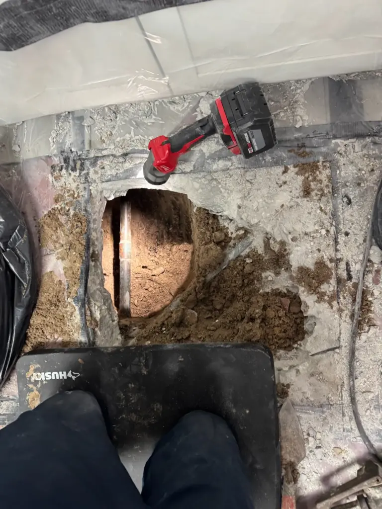 Technician kneeling on a pad at a hole dug through a slab, a copper pipe visible in the soil and a grinder beside it.