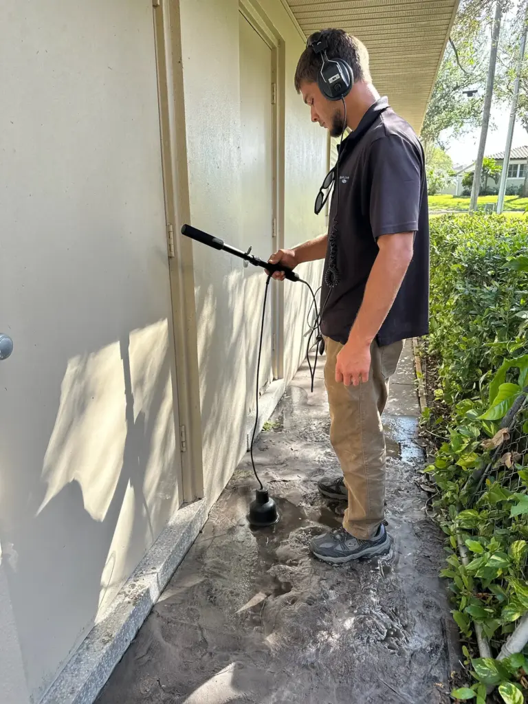 Technician wearing headphones holding an acoustic listening probe on a narrow walkway along a house exterior wall.