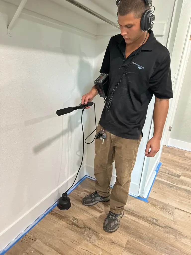 Technician wearing headphones holding an acoustic listening probe to a wood-look floor in an empty white room.