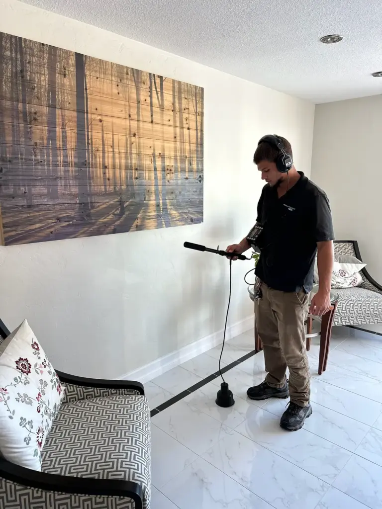 Technician wearing headphones holding an acoustic listening probe against a white tile floor in a living room.