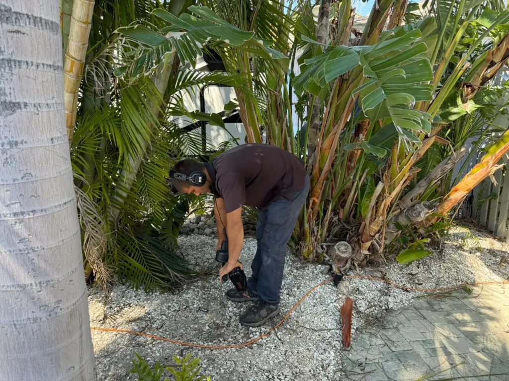 Technician wearing headphones bends over a shell rock landscape bed among palms, listening for a leak.
