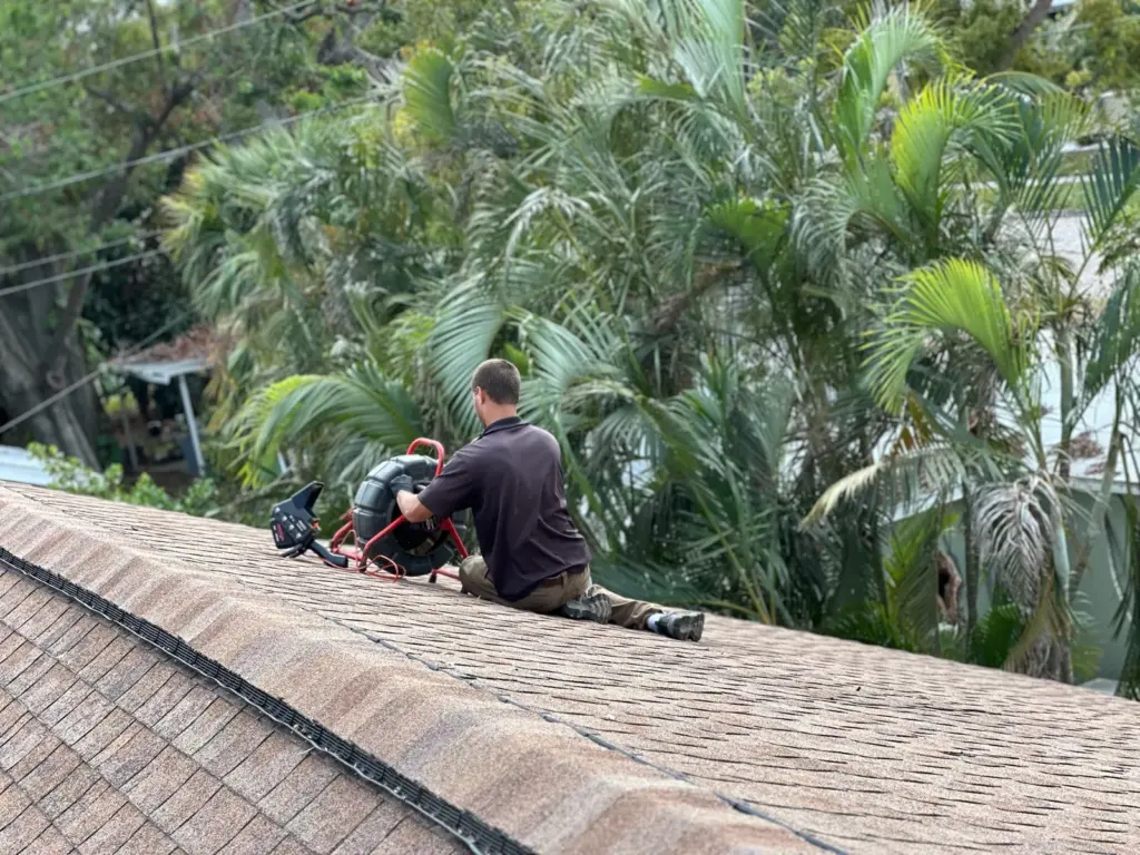 Technician kneeling near a roof ridge beside a red sewer camera reel, with palm trees behind him.