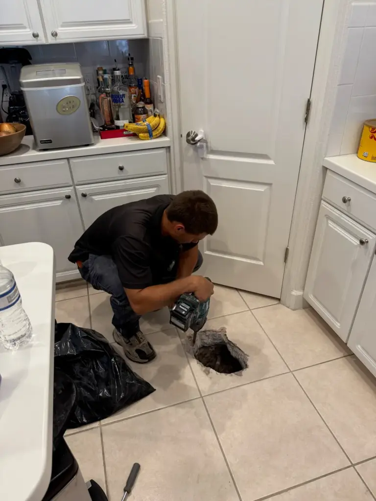 A technician crouches with a power tool beside a hole cut through a kitchen tile floor near a pantry door.