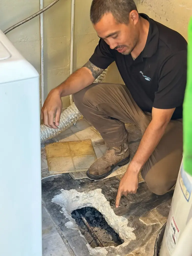 Technician crouched in a laundry room pointing down into an opening broken through the concrete floor.