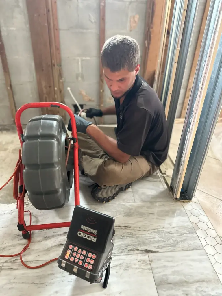 Technician seated on a tile floor feeding cable from a red sewer camera reel, with the camera monitor set beside him.
