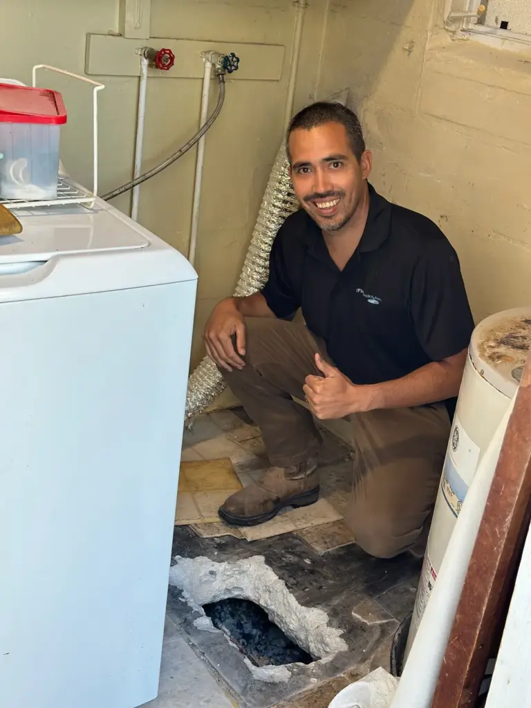 Smiling technician crouched in a laundry room giving a thumbs up beside an opening in the concrete floor.