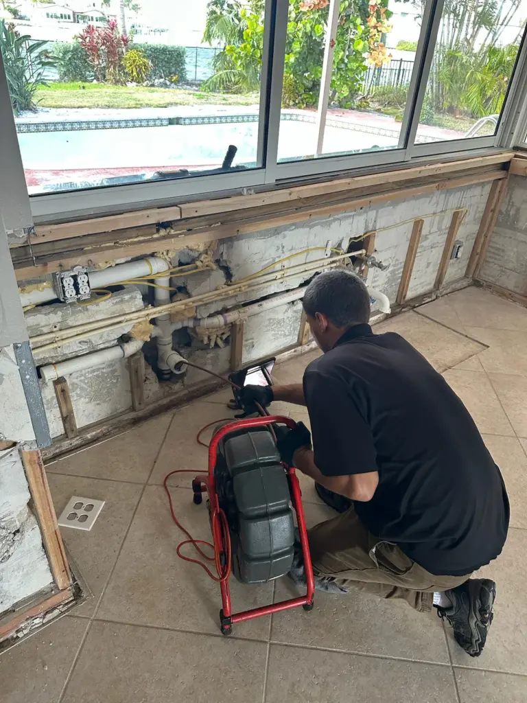 Technician kneels beside a red sewer camera reel at a gutted wall below a row of windows.