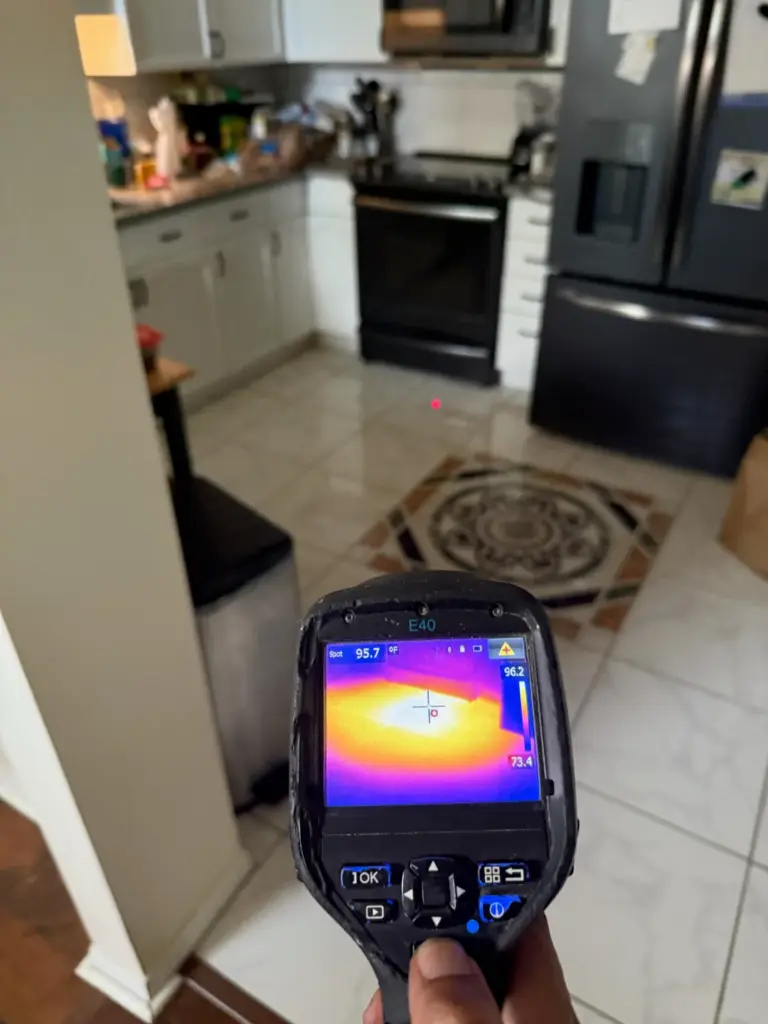 A thermal camera aimed at a kitchen tile floor shows a bright yellow and orange warm spot on its screen.
