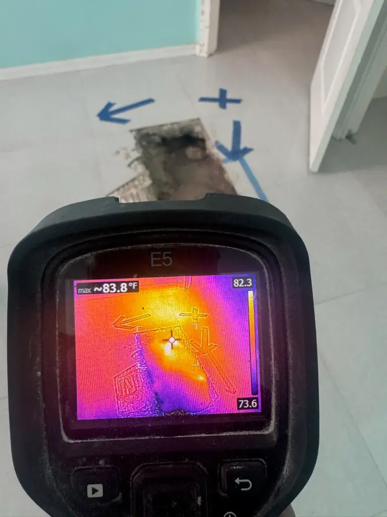 Thermal camera showing a bright warm spot, held beside a slab opening marked with blue tape arrows.