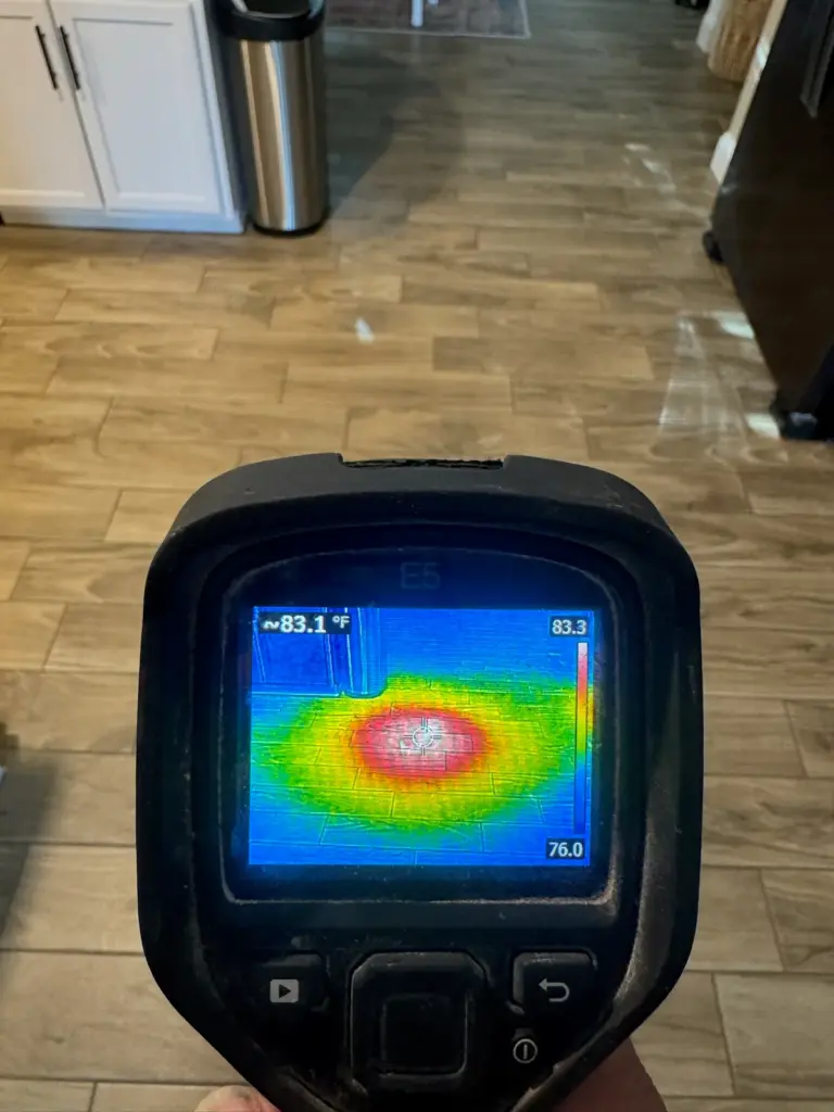 A thermal camera aimed at a wood-look tile kitchen floor, its screen showing a round red hot spot on cool blue.