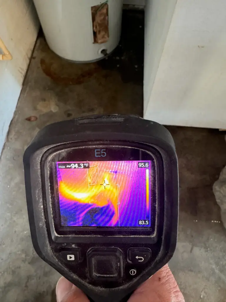 A thermal camera aimed at a stained concrete floor beside a water heater, its screen showing a bright hot streak.