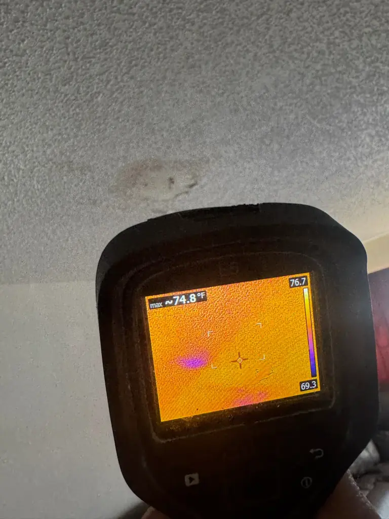 Thermal camera held up to a stained ceiling, its screen showing a small purple cool spot.