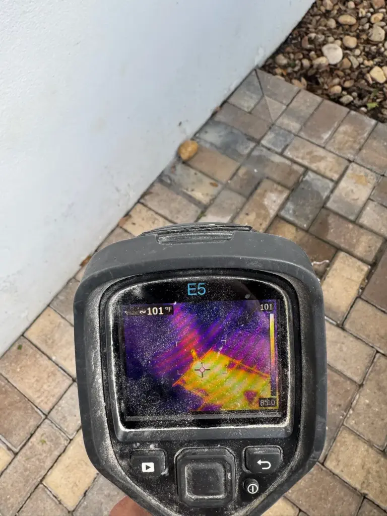 A handheld thermal camera showing a warm orange patch on brick pavers next to a white exterior wall.