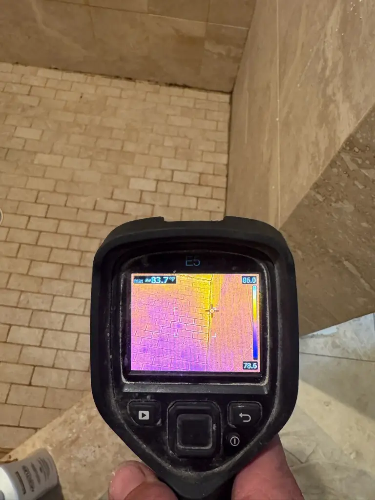 Thermal camera aimed at a tiled shower wall, its screen showing pink and orange tones.