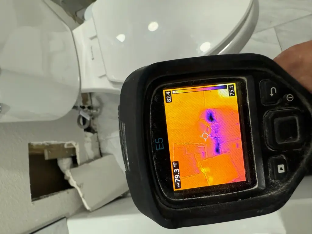Thermal camera beside a toilet, its screen showing purple cool streaks on an orange wall near an opening.