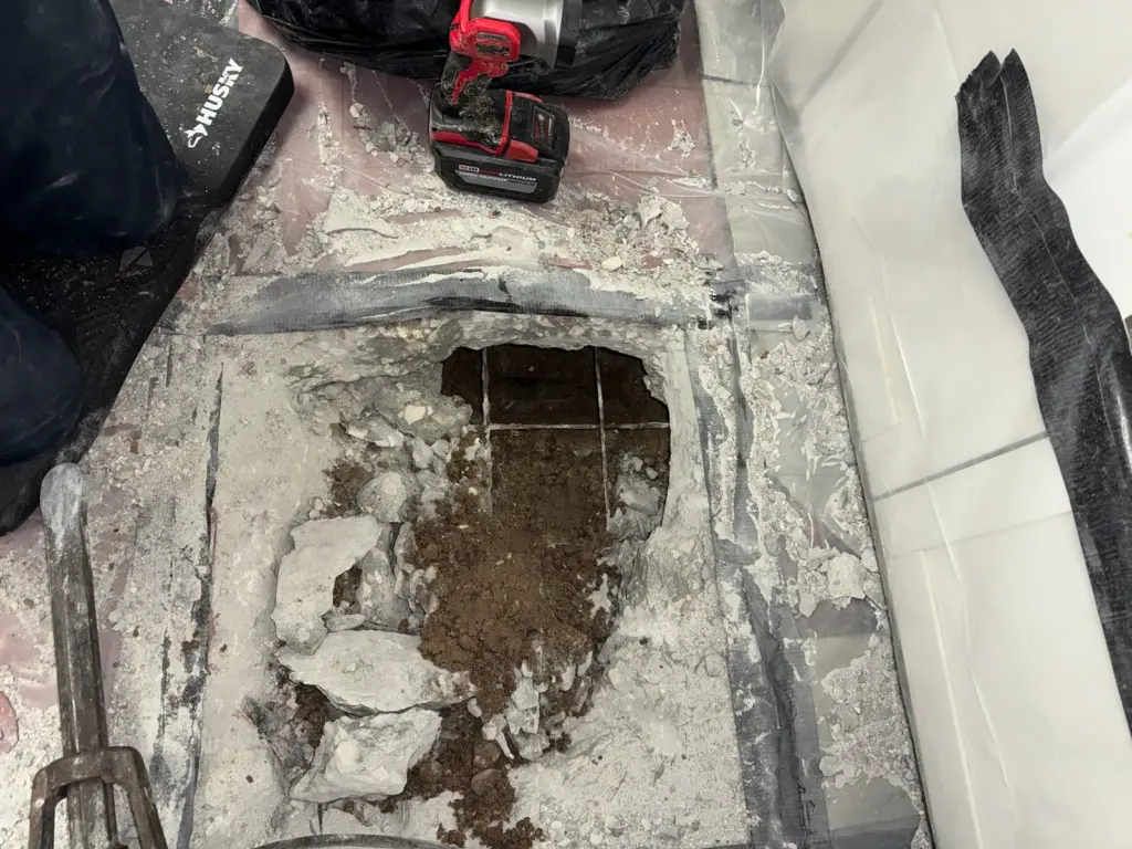 Tile and concrete slab broken out to expose soil below, with a red cordless tool and knee pad nearby.