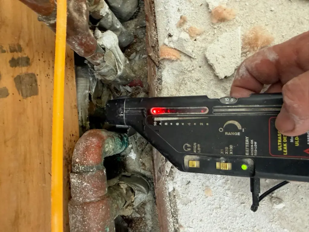 Hand holding an ultrasonic leak detector against corroded copper pipes inside an opened wall beside a wood stud.