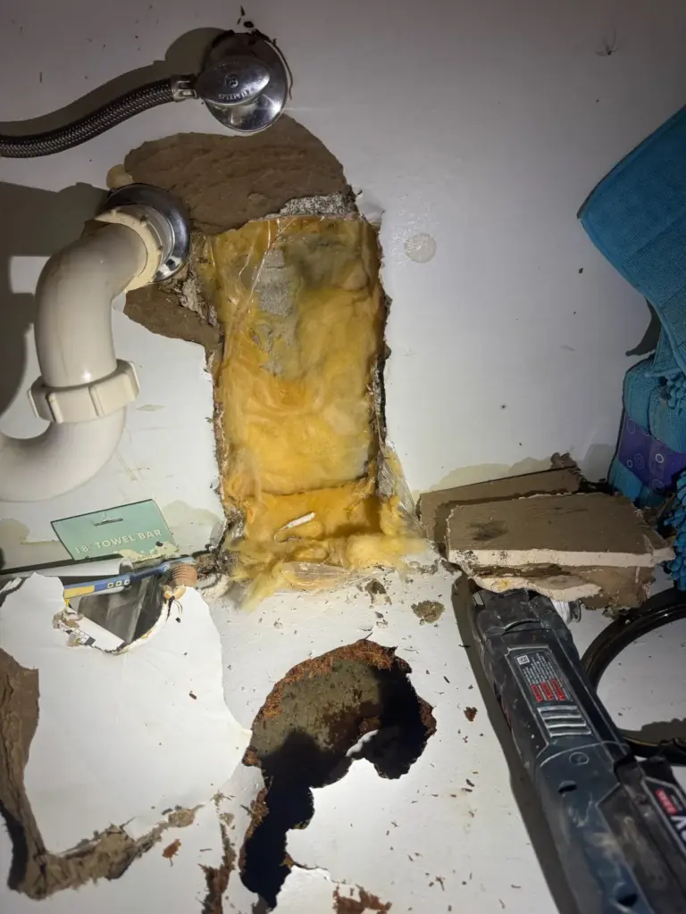 Drywall cut open behind a sink drain and supply line, exposing stained yellow insulation, with an oscillating tool below.