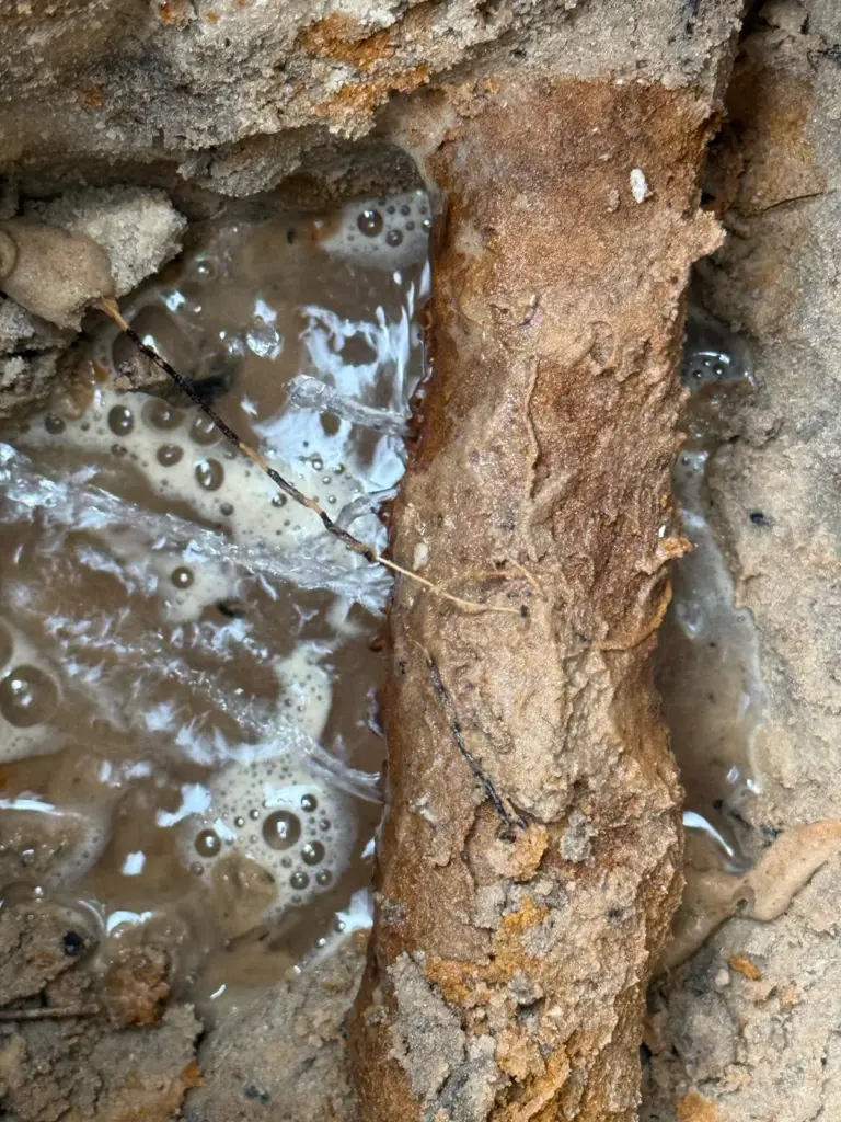 Close-up of water bubbling and foaming beside a heavily rusted metal pipe in a muddy excavation.