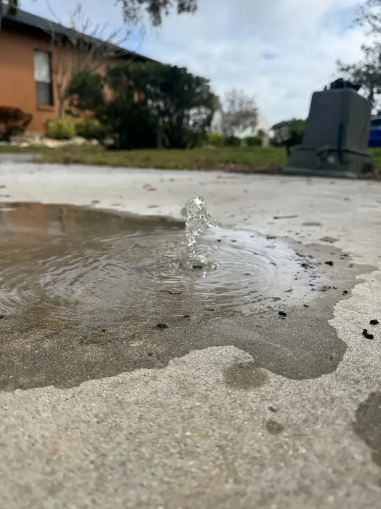 Water bubbling up through a crack in a concrete driveway, forming a spreading puddle.
