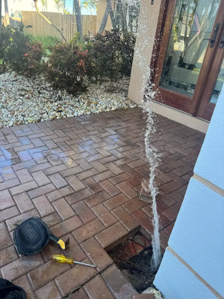 Water sprays high out of a small hole in a brick paver walkway beside a front entry, with a ground microphone nearby.