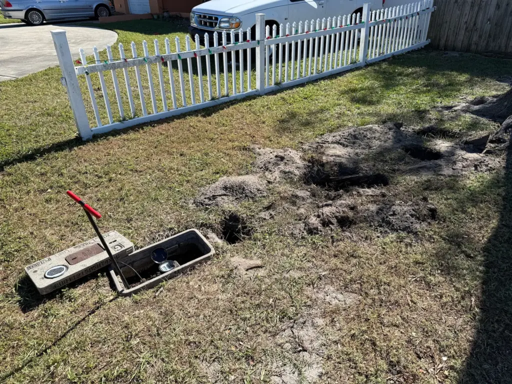Opened water meter box with a red-handled meter key, beside small holes dug in a lawn along a white picket fence.