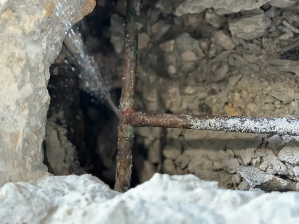 Fine spray of water shooting from a corroded copper pipe tee inside an opening broken through a concrete slab.
