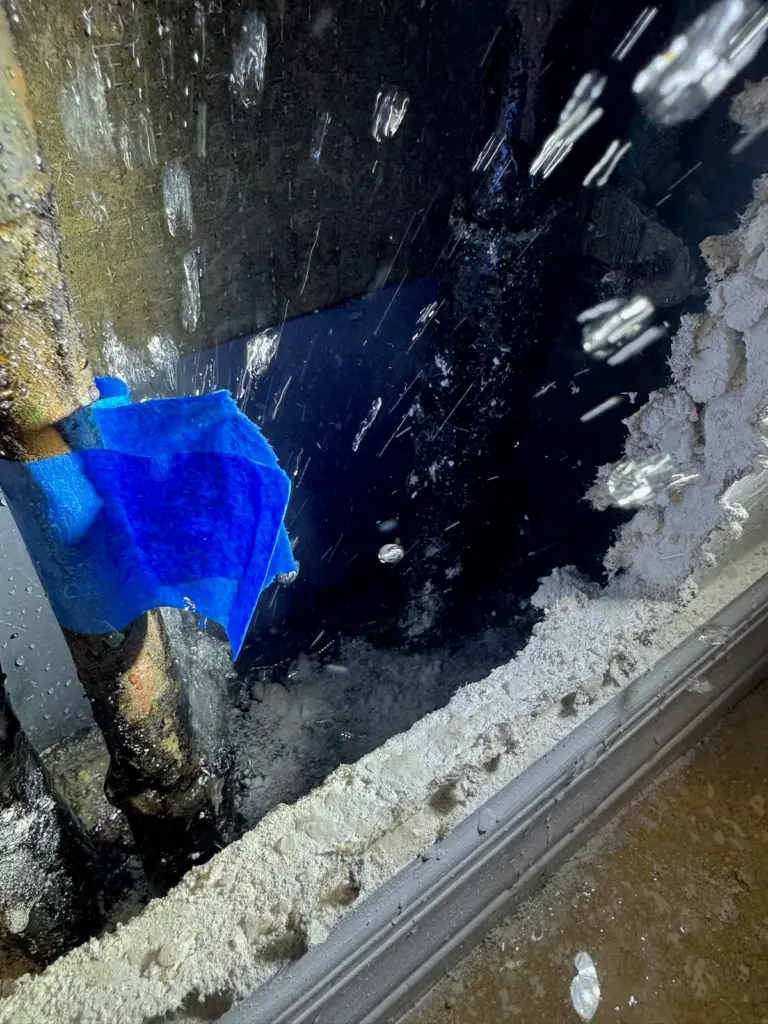 Water spraying from inside a wall opening, around a pipe partly wrapped in a blue cloth.