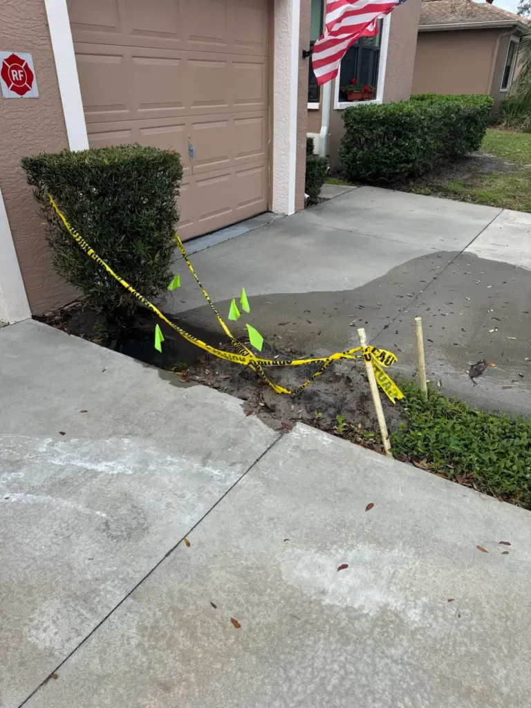 Water spreading across a concrete walkway from a garden bed marked with caution tape and green flags.