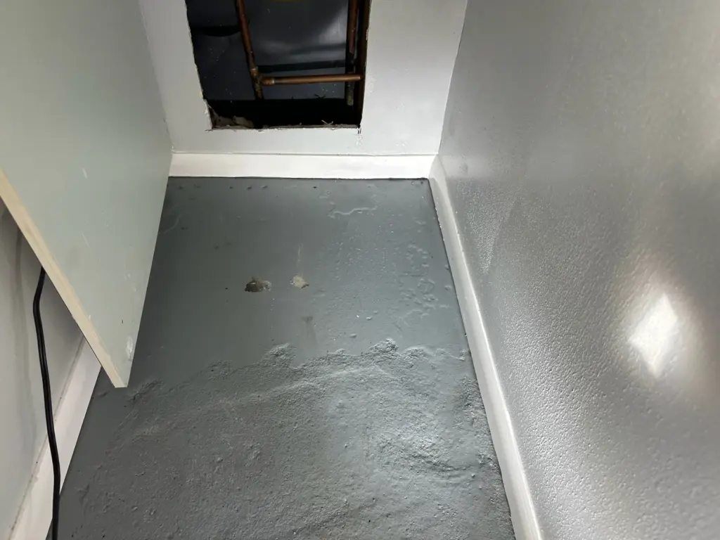 Wet gray painted concrete floor in a narrow closet below a wall opening that exposes copper water pipes.
