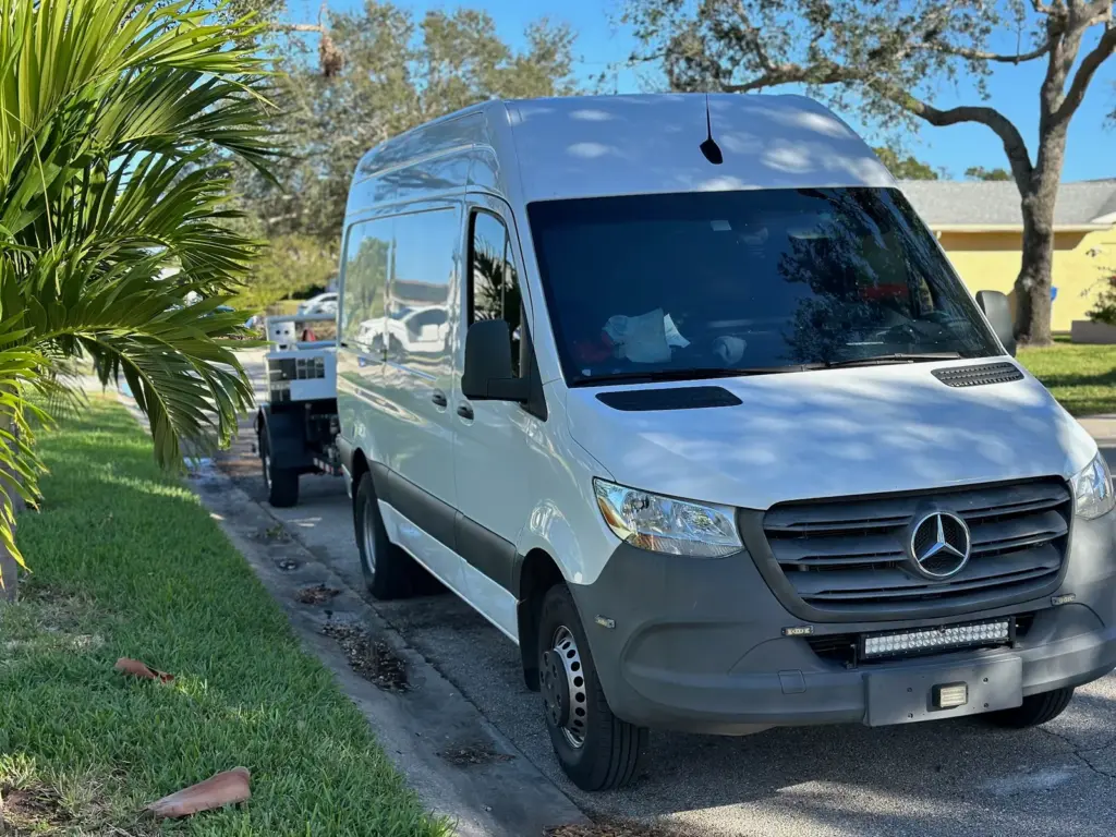 White Sprinter van parked along a grassy curb under palm fronds, a trailer hitched behind it.